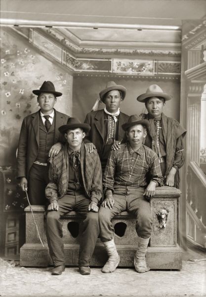Full-length studio portrait of five Ho-Chunk men posing in front of a painted backdrop. Two of the men are sitting on a prop stone wall, and three of the men are standing behind them. The men standing are, on the left, Will Blowsnake (One Who Stands and Strikes), (WoeGinNawGinKah), and two unidentified men. Sitting are Henry Badsoldier Stacy (MonNawPaeSheSheckKaw), left, and Harry Rave, a Nebraska Winnebago. All the men are wearing hats, and some of the men are wearing suit jackets, and vests. One man has a watch fob in his vest and a shawl over his shoulders.
