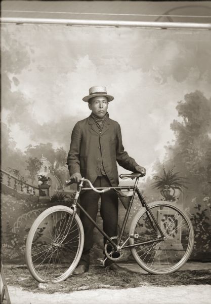 Full-length studio portrait of a young Ho-Chunk man, Jim Carriman, posing standing and holding a bicycle in front of a painted backdrop. He is wearing a hat, a scarf around his neck, a suit jacket, and trousers.