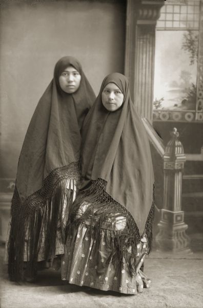 Kate Eagle (WoJayWinKah), left, and her sister Minnie (Edna) Eagle (DaScootchWinKah). Both women are wearing fringed shawls over their heads.