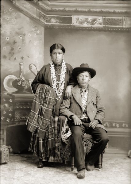 Full-length studio portrait in front of a painted backdrop of a Ho-Chunk couple, Belle Hall Greencrow (AhHooSkaWinKah), standing, and Henry Greencrow (CooNooZeeKah), sitting. She is wearing a dress, earrings, and long strands of beads, and is holding a plaid wool shawl. He is wearing a suit jacket, bandolier, tie, hat, and trousers.