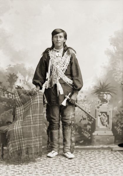 Full-length studio portrait of a young Ho-Chunk man, William Hindsley (Hensley) (CooNooChoNeeNikKah). He is holding a pipe tomahawk and is wearing bandoliers of bugle beads across his chest.