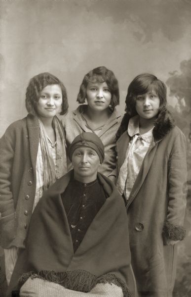 Studio portrait of a Ho-Chunk woman wearing a shawl and head scarf, posing sitting in front of her daughters, who are standing, in circa 1920s dress and short hair. Rachel Whitedeer Littlejohn (ENaKaHoNoKah), front, with daughters Florence Littlejohn Lamere (HoonchHeNooKah), left, Mary Littlejohn Fairbanks (WeHunKah), and Anne Littlejohn Lonetree.