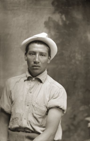 Waist-up studio portrait of a Ho-Chunk man, Raymond Pettibone, posing sitting and wearing a hat and a shirt with rolled-up sleeves in front of a painted backdrop.