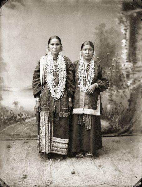 Full-length studio portrait in front of a painted backdrop of two unidentified Ho-Chunk women. The woman on the left is wearing a dress with many panels of narrow appliqué.