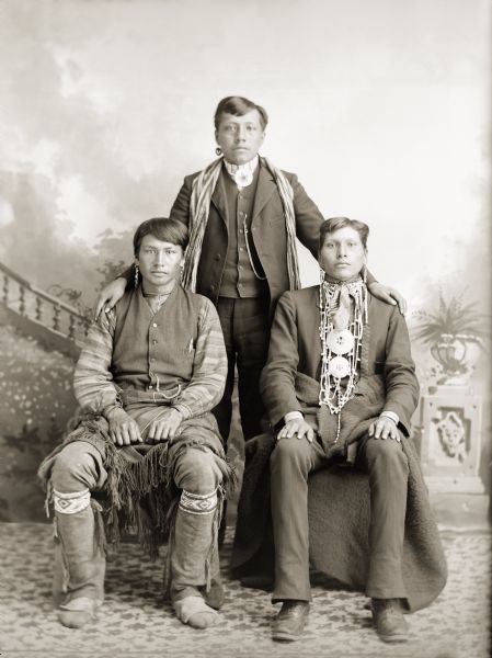Three unidentified men in a mix of contemporary and Ho-Chunk clothing. The man sitting on the right is wearing beads and a necklace made of large German silver medallions. The Ho-Chunk have always been metal workers, having made ornaments, armor, and functional objects from the lead and copper they mined, and later using trade metals.