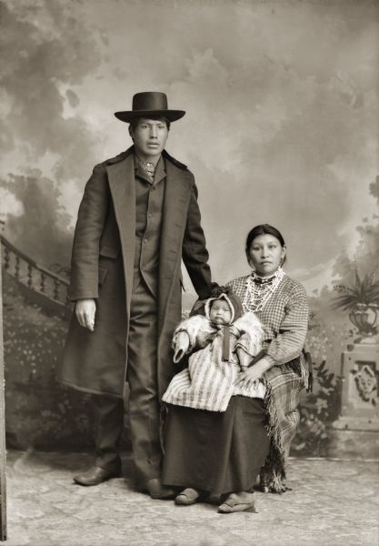 Full-length studio portrait of a Ho-Chunk man posing standing, and a Ho-Chunk woman posing sitting in front of a painted backdrop. The woman is holding a child in her lap. They are identified as George (Lyons) Lowe (AhHaZheeKah), who is dressed in a winter coat and hat and  his wife, Lena Nina Marie Decorah (Lyons) Lowe (AhHooSkaWinKah), and their infant son Martin (MaHisSkaKah). The Lowes were first listed on the census as Lyons, taking the name of a farmer they had worked for. The last name was later changed to Lowe, which is a shortened English version of their Indian name.