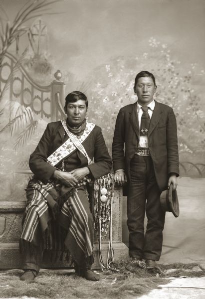 Full-length studio portrait of Joe Thunderking Lowe (King of Thunder) (WauKonJahHunkKah), sitting and wearing Ho-Chunk-style bandolier bags across his chest, an Indian trade blanket on his lap, and Ho-Chunk moccasins. He is leaning on a pedestal of a prop stone wall that is draped with bandoliers of beads, hair pipe bone, and silver conches. David Davis W. Decorra (NeZhooLaChaHeKah), on the right, is dressed in contemporary clothing that has been accented with a Ho-Chunk beaded belt and a tie decorated with German silver brooches (heapoke).