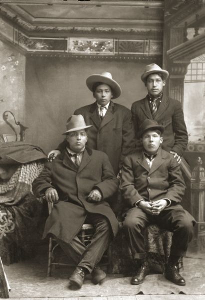 Full-length studio portrait in front of a painted backdrop of two Ho-Chunk men posing sitting in front of two Ho-Chunk men posing standing. All the men are wearing hats and neckties, and wearing suit jackets except the man sittingon the left. Identified as George Lonetree (HoonchXeDaGah), standing left, and Harry Funmaker (HaGaChaCooKah); seated are Fred Peter Wildrice Bearchief (WaGeSeNaPeKah), left, and Julius Whitedog (ChakShepWasKaHeKah).