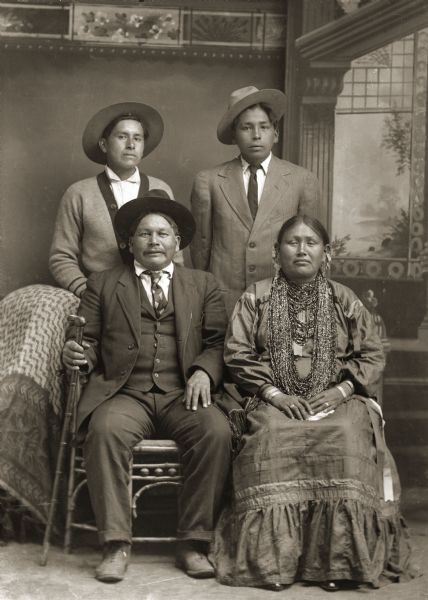Studio portrait of group of people posing in front of a painted backdrop. A Ho-Chunk man is sitting on the left wearing a suit, necktie, and hat, and is holding a cane in his right hand. A Ho-Chunk woman is sitting on the right who is wearing a light-colored dress, several necklaces, earrings, and file bracelets. Behind the man and woman are two Ho-Chunk men standing who are wearing hats. The man on the left is wearing a sweater, and the man on the right is wearing a suit and necktie. They are identified as Hugh Lonetree (CooNooKah), standing left, and George Lonetree (HoonchXeDaGah); seated are Alec (Alex) Lonetree (NaENeeKeeKah) and his wife, Lucy Emerson Brown, a Winnebago from Nebraska.