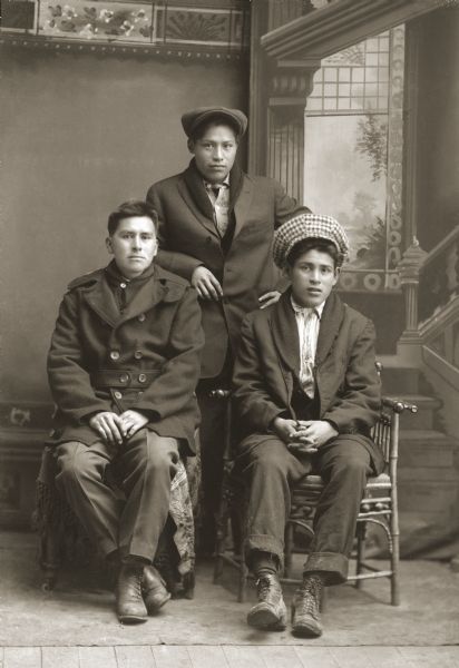 Studio portrait in front of a painted backdrop of two young Ho-Chunk men posing sitting in front of another young Ho-Chunk man posing standing. The man on the left is wearing a pea coat and cap, the man in the center is wearing a suit and cap, and the man on the right is wearing a suit coat and hat. They are identified as, from left to right, Hugh Lonetree (CooNooKah), Jesse Stacy (CooNooKah), and Alliston Winneshiek (MoNaKaKah).
