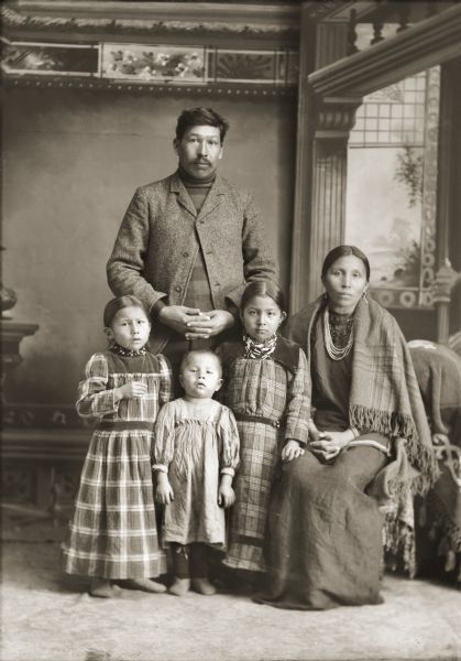 Full-length studio portrait in front of a painted backdrop of a Ho-Chunk family. The man is posing standing behind the group and is wearing a suit coat. In front of him are three children, all wearing dresses. To the right of the girls is a Ho-Chunk woman posing sitting and wearing earrings, necklaces, and a shawl around her shoulders. Identified as George (Lyons) Lowe (AhHaZheeKah) with his wife, Lena Nina Marie Decorah (Lyons) Lowe (AhHooSkaWinKah) and family. Standing in front of George are their children, Lydia Lowe, Daniel Lowe, and Bessie Lowe.
