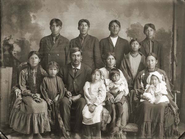 Studio portrait of a large Ho-Chunk group posing in front of a painted backdrop, including five men, three women, three girls, and two smaller children. The family of Joe Thunderking Lowe (King of Thunder) (WauKonJahHunkKah). Standing in back from left to right are Gilbert Thunderking Lowe (WaConChaHoNoNeeKah), Albert Thunderking Lowe (ChaShepSkaKah), Fred Kingswan (MaHeNoGinKah), Annie Thunderking Lowe (WaNukScotchEWinKah), and Sam Thunderking Lowe (HoChumpHoNikKah). In the front row from left to right are Lucy Thunderking Lowe Yellowbank (MaHiskaWinKah), Minnie Thunderking Lowe (ENooKahHooNooKah), Joe Thunderking, Dora Thunderking Lowe (AkSeKahHoNoKah), Mary Decorah Thunderking (KaNooKayWinKah), Theodore Thunderking Lowe (WaConChaHoNoNikKah), sitting on his mother Mary’s lap, and Alice Thunderking Lowe Kingswan (MaHayPaSayWinKah) holding Violet Kingswan (WeGoSeeDaWinKah).