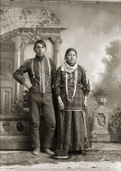 Full-length studio portrait of a Ho-Chunk man and woman posing standing in front of a prop stone wall and painted backdrop. A hat is on the ground in front of them. The man is wearing trousers with suspenders, a beaded necklace, and ribbons. The woman is wearing earrings, bracelets, and beaded necklaces. They are identified as Edward Funmaker (WaGeSeNaPeKah) and his wife, Mamie (Minnie) Bearchief Funmaker (HoHumpCheKaRaWinKah).