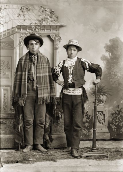 Full-length studio portrait of two young Ho-Chunk men standing in front of a prop stone wall and a painted backdrop. On the right is Tom Thunderchief, (son of Child of Thunder Winneshiek), who is wearing a Ho-Chunk beaded shirt, belt, and a hat with a beaded hat band.