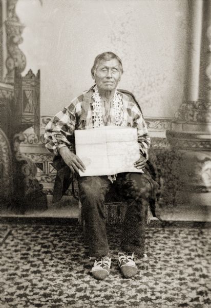 Full-length studio portrait of an elderly Ho-Chunk man posing sitting in front of a painted backdrop. Green Grass (HaWinChoKah) is wearing floral beaded Ho-Chunk moccasins, and is holding a map of Jackson County that shows his 40-acre allotment.