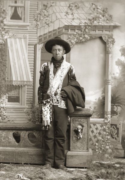 Full-length studio portrait of Frank Washington Lincoln (ChakShepKonNeKah), dressed in the style of a single man with crossed bandolier bags. He is wearing a hat and regalia, including earrings, and beaded necklaces. He is standing in front of a low, prop stone wall in front of a painted backdrop.