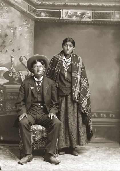 Full-length studio portrait in front of a painted backdrop of a Ho-Chunk man and woman. Identified as Frank Washington Lincoln (ChakShepKonNeKah) and his first wife, Betty Otter Lincoln (NaChoPinWinKah). Frank is posing sitting and is wearing earrings, as well as a suit jacket, vest with watch fob, tie, and a hat. Betty is standing next to him and is wearing earrings, beads and a plaid shawl over a long skirt.
