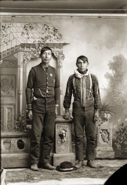 Full-length studio portrait of two Ho-Chunk men posing standing in front of a painted backdrop near a prop stone wall. A hat is on the ground between them. The taller man on the left, identified as Albert Thunderking Lowe (ChaShepSkaKah), has a handgun in his pocket and is wearing a badge on his vest. The man on the right, Edward Funmaker (WaGeSeNaPeKah), is wearing a flannel shirt with two ribbons (zeeniba) sewn down the front.
