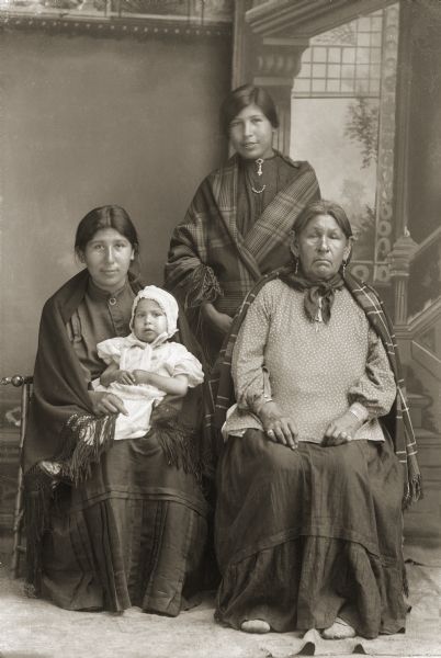 Studio group portrait in front of a painted backdrop. Lucy Thunderking Lowe Yellowbank (MaHiskaWinKah), sitting on the left, is holding her nephew David Neil Lincoln Jr. (WaWaHasKah), with her sister, and mother of David, Annie Thunderking Lowe Lincoln (WaNukScotchEWinKah), standing and Sarah Half-a-Sack Longmarsh (NeeZhooJhotchKayWinKah)(WaNeekSootchEWinKah) sitting on the right.