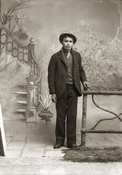 Full-length studio portrait in front of a painted backdrop of a young Ho-Chunk man posing standing near a prop fence. He is wearing a suit jacket, vest with watch fob, a scarf around his neck, trousers, and a hat. He is identified as Frank Yellowfeather Climer (HaGaKaw). Frank and Emma E. Climer (ENooKaStaLaKah) are the parents of Susie and Bertha Climer.