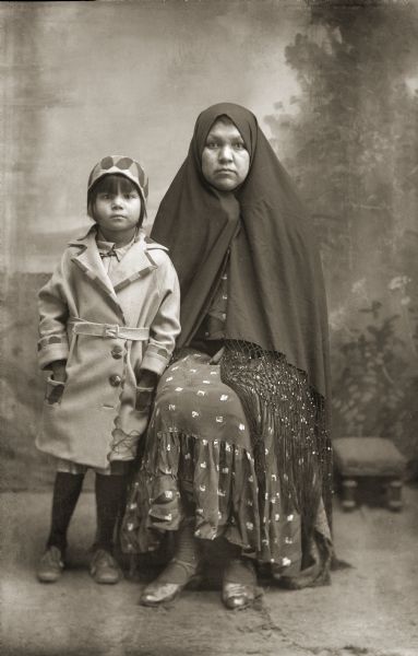 Full-length studio portrait of a Ho-Chunk woman and girl in front of a painted backdrop. The woman is posing sitting and wearing a shawl over her head, and the girl is posing standing and wearing a modern coat and matching hat. Identified as Agnes Payer Climer, and her daughter Juanita June Climer.