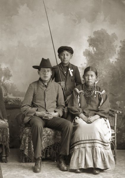 Full-length studio portrait of a young Ho-Chunk man and woman posing sitting in front of a Ho-Chunk boy standing in front of a painted backdrop. The boy, James or George Swan, is holding a fishing pole. Harry Littlesnake (HaKaChoMonEKah) and Annie Youngswan Snake (WaKaChaMaNeeKah).