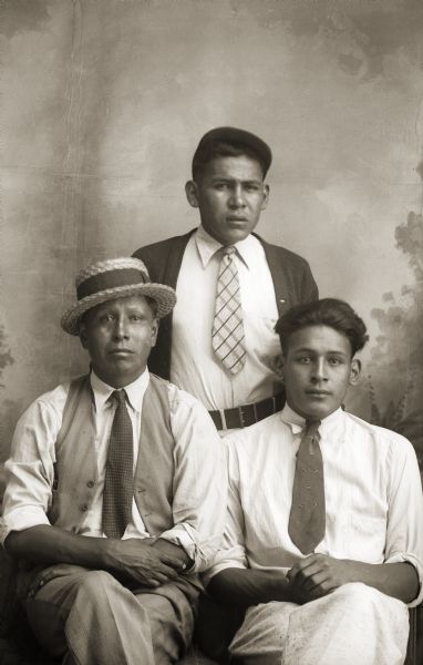 Studio group portrait of three Ho-Chunk men posing in front of a painted backdrop. Two men are all wearing neckties. Mark Henry is standing in the center behind Fred Kingswan (MaHeNoGinKah), left, and his brother Elias Henry. The Henry boys were from Nebraska.
