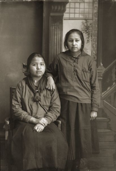 Three-quarer length studio portrait in front of a painted backdrop of two young Ho-Chunk women wearing Middy blouses. Lucy Whitebear (HoDaHooKah) is sitting next to Mary (Pinkah) Lewis (KeSaWinKah) who is standing.