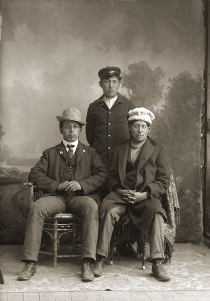 Full-length studio portrait in front of a painted backdrop of three Ho-Chunk men. Two men are sitting in front of a man standing behind them in the center. They are all wearing hats. Frank Lewis (HaNaCheNaCooNeeKah) is standing behind Fred Kingswan (MaHeNoGinKah), left, and John Swallow (MonKeSaKaHepKah).