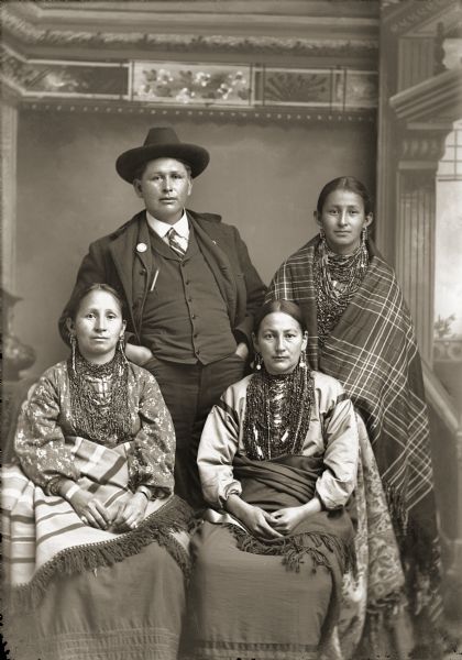 Studio portrait in front of a painted backdrop of a group of four Ho-Chunk Indians. Little Sam (HePaHeKah), a Ho-Chunk man, is posing standing and wearing a suit, necktie, and hat. His wife Rachel (White Deer) Little Sam stands next to him wearing necklaces and earrings, and is wrapped in a plaid shawl. Margaret Whitedeer Little Sam (NauChoPinWinKah) is seated to the left of Sarah French (Ellen Davis) (AhHooChoWinKah). They are posed sitting wearing necklaces and earrings, and holding shawls/blankets on their laps.