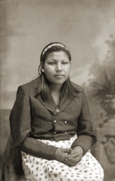 Studio portrait in front of a painted backdrop of a young Ho-Chunk woman posing sitting with her hands clasped. She is wearing a beaded head band, a dark blouse, and a light-colored printed skirt, and is identified as Dorothy Greengrass, daughter of Robert Greengrass (HoNutchNaCooMeeKah) and Dora Winneshiek (HoHaWinKah).