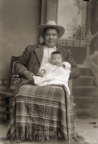 Studio portrait in front of a painted backdrop of a Ho-Chunk man posing sitting and wearing a suit and hat, and with a shawl on his lap. Jim Whitebear (HoonchSkaKah) is holding a baby boy, possibly his son Dan Whitebear (HeNukKah).