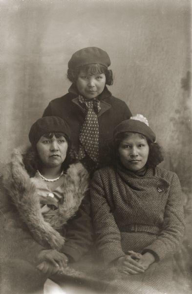 Three-quarter length studio portrait of three young Ho-Chunk women. One of the women is posing standing in the center behind the other two woman posing sitting. They are all wearing modern dress and hats. Bertha Climer is standing behind her sister-in-law, Agnes Payer Climer, on the left, and her sister Susie Climer on the right. Agnes was married to Bertha and Susie’s brother, Thor.
