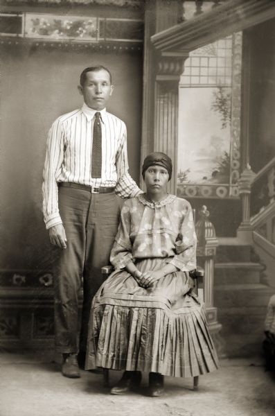 Full-length studio portrait of a Ho-Chunk man and his wife posing in front of a painted backdrop. The man is standing and wearing a tie, and the woman is sitting. They are both wearing contemporary dress. Felix White-Wilson (PawNeeKaw) of Nebraska is with his second wife, Agnes Eagle (HaHumpAWinKah).