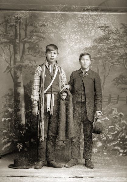 Full-length studio portrait of two young Ho-Chunk men. The man on the left, Frank C. Thunder (Hoonk Ta Ka Kah), is wearing streamers over his shoulders and holding a shawl/blanket. The man on the right, Edward Greengrass (Che Win Che Kay Ray He Kah), is wearing a suit jacket and is holding a hat by his side, while resting his hand on the blanket held by the other man. In the background is a painted backdrop.