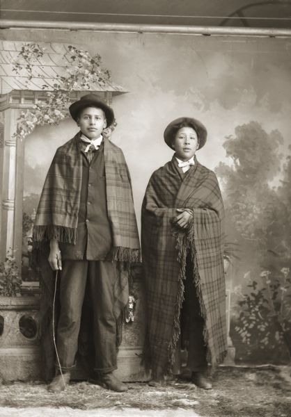Full-length studio portrait in front of a painted backdrop of two young Ho-Chunk men posing standing near a prop stone wall. Charlie Greengrass (HoeHumpCheeKayRayHeKah) is standing to the left of Will Thunder (NaNikSayWaHeKah).