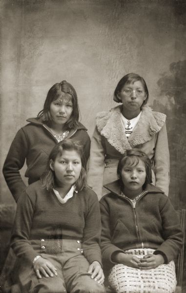 Studio portrait in front of a painted backdrop of two adolescent Ho-Chunk women posing standing behind two adolescent Ho-Chunk women posing sitting. They are all wearing contemporary clothing and have short hair. Seated are Dorothy Greengrass, left, and Edna Climer. Standing are Jennie Climer, left, and Ollie May Sine. Jennie and Edna were twins.
