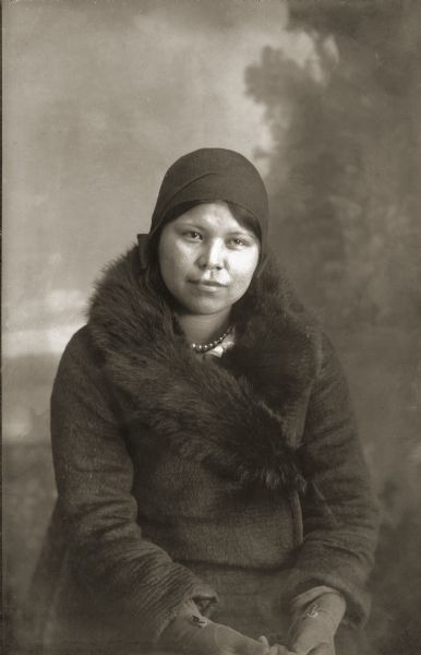 Studio portrait in front of a painted backdrop of a Ho-Chunk woman posing sitting. She is wearing a dark coat with a fur-lined collar, gloves, cloche hat, and a pearl necklace. She is identified as Emma Climer, the mother of Susie.