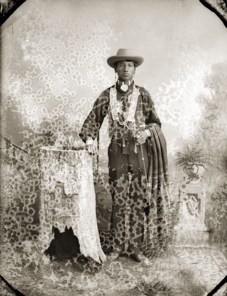 Full-length studio portrait of (Hoonk Ha Ga Kah) (Son of [Wa Con Cha Kah[ John Thunder aka Dr. Thunder and [We Hon Pe Kaw] Lucy Bear, Thunder). He is leaning on a stump used as a prop in Van Schaick’s studio and is adorned with earrings and a choker. A blanket is draped over his arm. In the background is a painted backdrop.