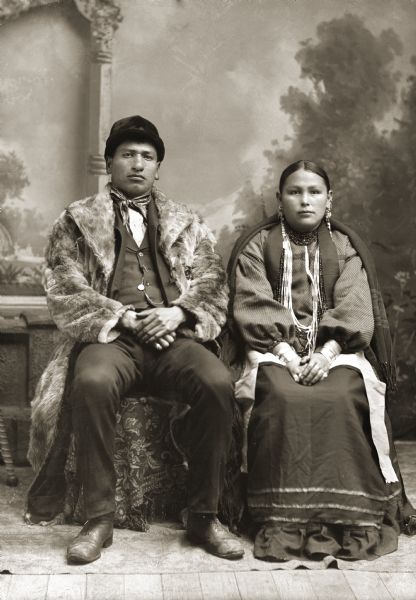 Full-length studio portrait of a Ho-Chunk man and his wife posing sitting in front of a painted backdrop. He is wearing a fur coat, suit jacket, vest with watch fob, and hat, and she has on a wool shawl, necklaces, and long earrings. They are identified as Thomas Thunder (HoonkHaGaKah) and his wife, Florence Littlesoldier Wallace Thunder (MauKonNee WinKah), who lived at Hunter’s Ridge.