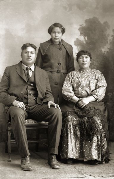 Full-length studio portrait of a Ho-Chunk family. Sitting (l to r) Will Thunder (NaKikSayWaHeKah) (son of [WaConChaKah] John Thunder aka Dr. Thunder and [WeHonPeKaw] Lucy Bear, Thunder) and his wife, Mary White, Greencloud, Thunder. Standing behind them is their daughter Ivy (Kate Greencloud) Thunder. In the background is a painted backdrop.