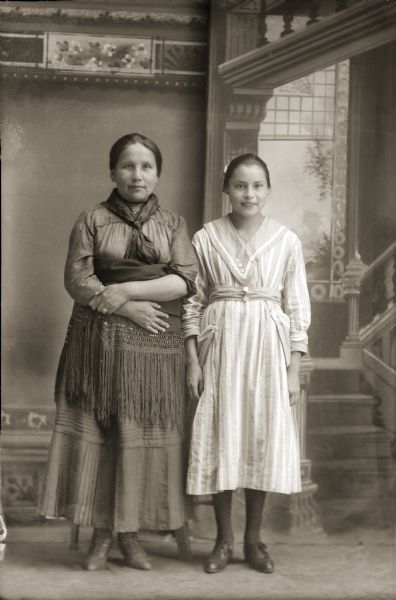 Full-length studio portrait in front of a painted backdrop of two Ho-Chunk women posing standing. The older woman on the left is holding a fringed shawl around her torso, and the younger woman on the right is wearing a light-colored striped dress. Kate Thunder Miner (WaRoSheSepEWinKah) standing next to Hilda Alice Stacy Funmaker (HoWaChoNeWinKah).