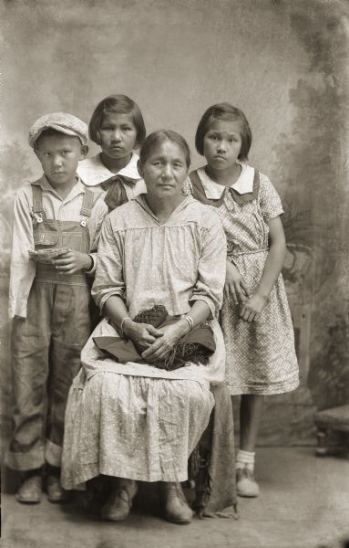 Studio portrait in front of a painted backdrop of a Ho-Chunk woman posing sitting and wearing a light-colored dress with a fringed shawl on her lap. She is in front of two Ho-Chunk girls posing standing behind her, and who are wearing the same style of light-colored dress. A Ho-Chunk boy is posing standing on the left, wearing overalls and a cap and holding something in his left hand. They are identified as Lucy Wilson Blackhawk (HoChunkEWinKah), seated, with, left to right, Donald (Dun) Blackhawk, Margaret Blackhawk, and Virginia Blackhawk.