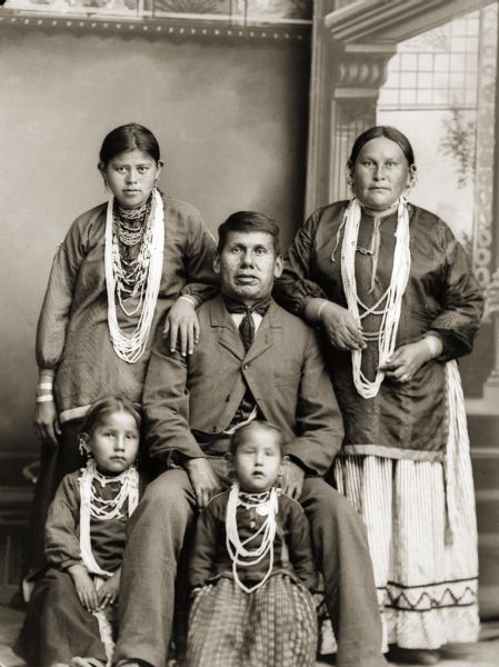 Studio portrait in front of a painted backdrop of a Ho-Chunk man posing sitting in the center wearing a suit and necktie, behind two Ho-Chunk girls posing sitting on the floor who are wearing several necklaces and earrings. Two women are posing standing behind the man and girls, and are wearing several necklaces and earrings. William Thunder (WaConChaHaTeKah) is seated to the left of his wife Mary Mollie Prophet Thunder (HoWaChoNeWinKah). Their children are seated in front of them, Kate Thunder (WaRoSheSepEWinKah), left, and Lucy Thunder (ENooKahKah). The young woman resting her hand on William’s shoulder is possibly identified as x’ax it nelic, her Indian name in the old Ho-Chunk writing system.
