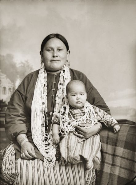 Studio portrait of a Ho-Chunk woman posing sitting in front of a painted backdrop wearing several necklaces, earrings, and a striped skirt. In her lap she is holding an infant in a striped dress who is wearing several necklaces. Mary Mollie Prophet Thunder (HoWaChoNeWinKah) holding her daughter Kate Thunder (WaRoSheSepEWinKah).