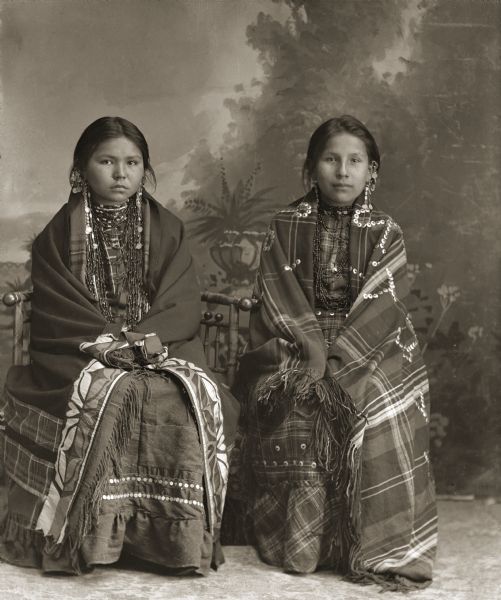Full-length studio portrait of two Ho-Chunk girls posing sitting in front of a painted backdrop. Caroline Mary Decorah Blackdeer (CheHeTeChaWinKah), left, is wearing a silk appliqué blanket (wai zeenibawoore), and Kate Thunder Miner (WaRoSheSepEWinKah) is wearing a shawl (wai) embellished with silver brooches (hiiwapox).
