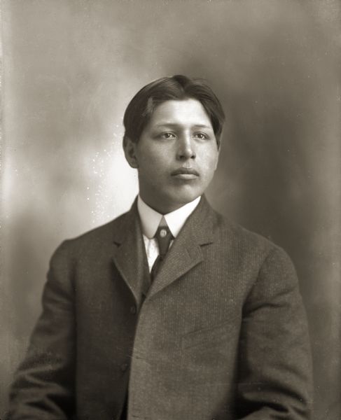 Waist-up studio portrait of a Ho-Chunk man with short hair posing sitting. He is wearing a suit coat and necktie, and is identified as Fred Kingswan (MaHeNoGinKah).