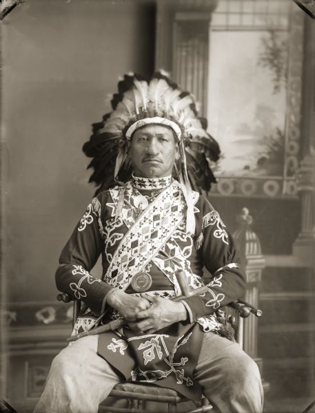 Three-quarter length studio portrait of Thomas Thunder (Hoonk Ha Ga Kah) (Son of [Wa Con Cha Kah] John Thunder aka Dr. Thunder and [We Hon Pe Kaw] Lucy Bear, Thunder). He is wearing elaborate Ho-Chunk regalia including eagle feather bonnet, floral beaded shirt and breechcloth, loom-beaded bandolier bags, choker and an Indian Peace Medal. He is holding a calumet pipe. In the background is a painted backdrop.