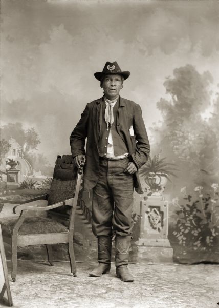 Full-length studio portrait of a Ho-Chunk man posing standing next to a chair, identified as Homer Snake, who is wearing a hat with a GAR emblem, signifying he is a Civil War veteran. Snake served as a private with Company A of the Omaha Scouts, a Civil War unit composed of six white officers and seventy-two Ho-Chunk soldiers.