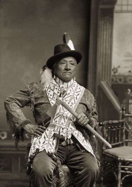 Three-quarter length studio portrait of an elderly Ho-Chunk man Henry Rice Hill (SanJanMonEKah), posing in front of a painted backdrop.  Henry was a private in Company A of the Omaha Scouts during the Civil War. He is wearing a deer bone choker around his neck and two Ho-Chunk bandolier bags across his chest, and is holding a pipe tomahawk. The golden eagle tail feather in his hat signifies warrior or veteran status.